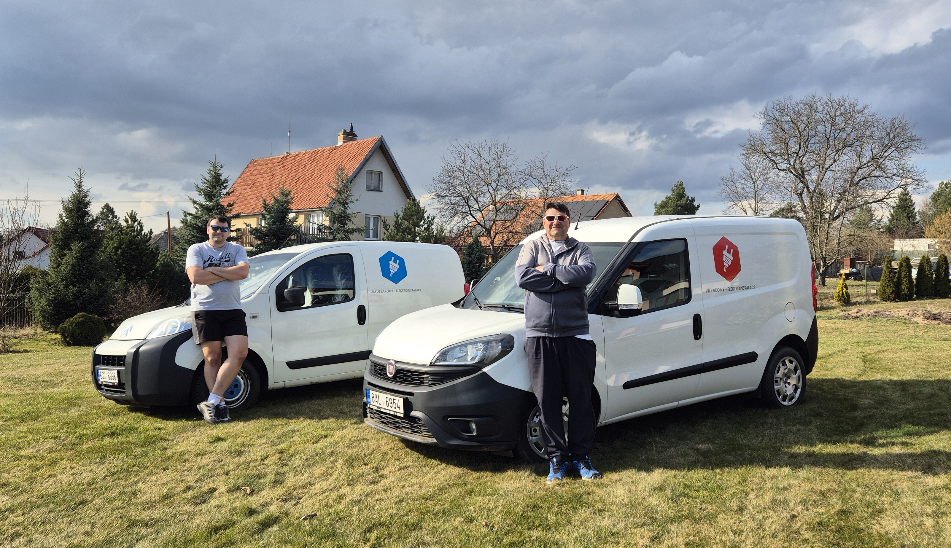 Dva elektrikáři z Elektroinstalace Lakomý stojí před dvěma firemními dodávkami s logem firmy na klidné zahradě rodinného domu. Fotografováno naležato, zepředu a z mírného odstupu tak, aby byly dobře vidět vozy, lidé i okolní zástavba. Snímek působí přátelsky, důvěryhodně a reprezentuje firmu na úvodní stránce.
